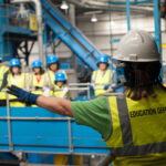 An education officer pointing out recycling machines to a group of people. All are wearing hi vis vests and safety helmets.