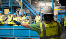 An education officer pointing out recycling machines to a group of people. All are wearing hi vis vests and safety helmets.