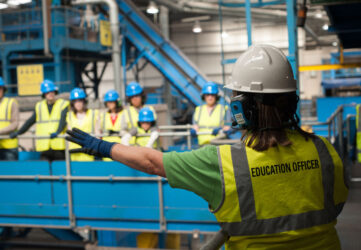 An education officer pointing out recycling machines to a group of people. All are wearing hi vis vests and safety helmets.