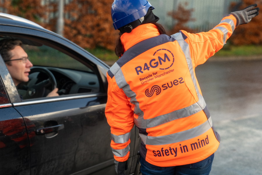Photo of someone who works at a recycling centre in greater manchester for recycle for greater manchester. They are pointing to direct someone driving a car to the right area of the recycling centre.